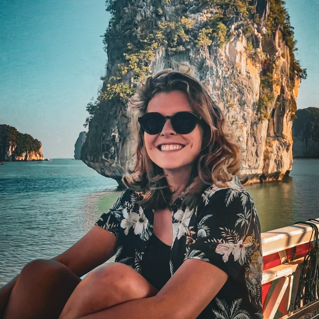 A smiling person wearing sunglasses and a floral shirt, sitting on a boat with scenic rocky cliffs and calm waters in the background.